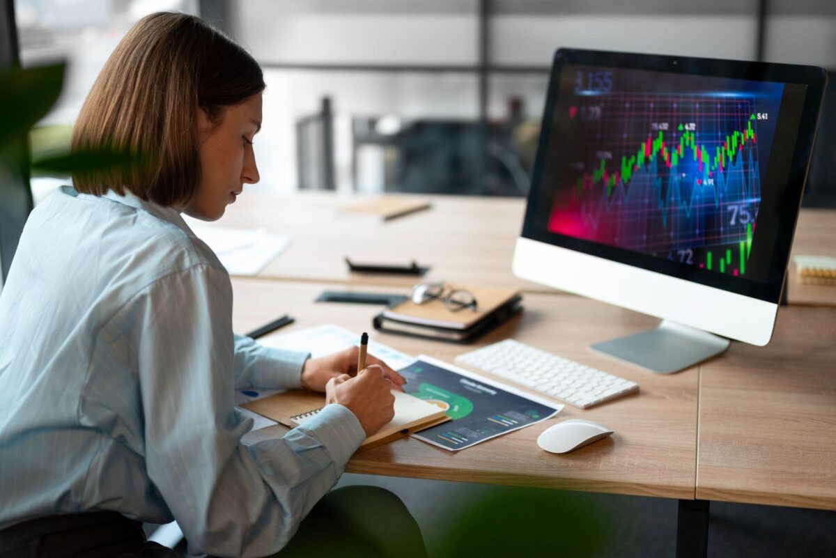 A lady writing on a note pad with a stock trading screen in front of her to illustrate the story